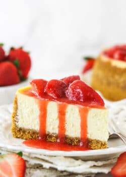 A slice of strawberry cheesecake on a plate with a fork near fresh strawberries and a full strawberry cheesecake.